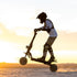 A person wearing a helmet and protective gear rides a black Kaboo electric scooter on a flat desert surface at sunset, with the scooter performing a wheelie.