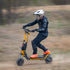 A person riding an electric scooter on a dirt path, wearing a white helmet, black protective gear, and bright orange shoes.